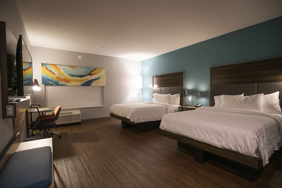 A hotel room with two beds, a desk with a chair, a TV, and artwork on the wall.