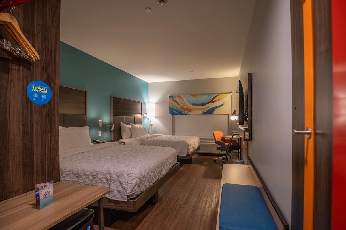 A hotel room with two beds, a desk, a chair, a desk lamp, and a painting on the wall.