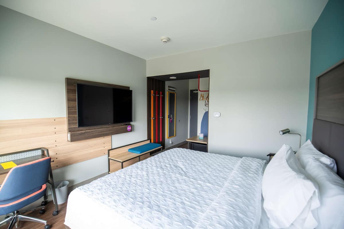 A modern hotel room with a large bed, a desk with a chair, a television mounted on the wall, and a wardrobe.