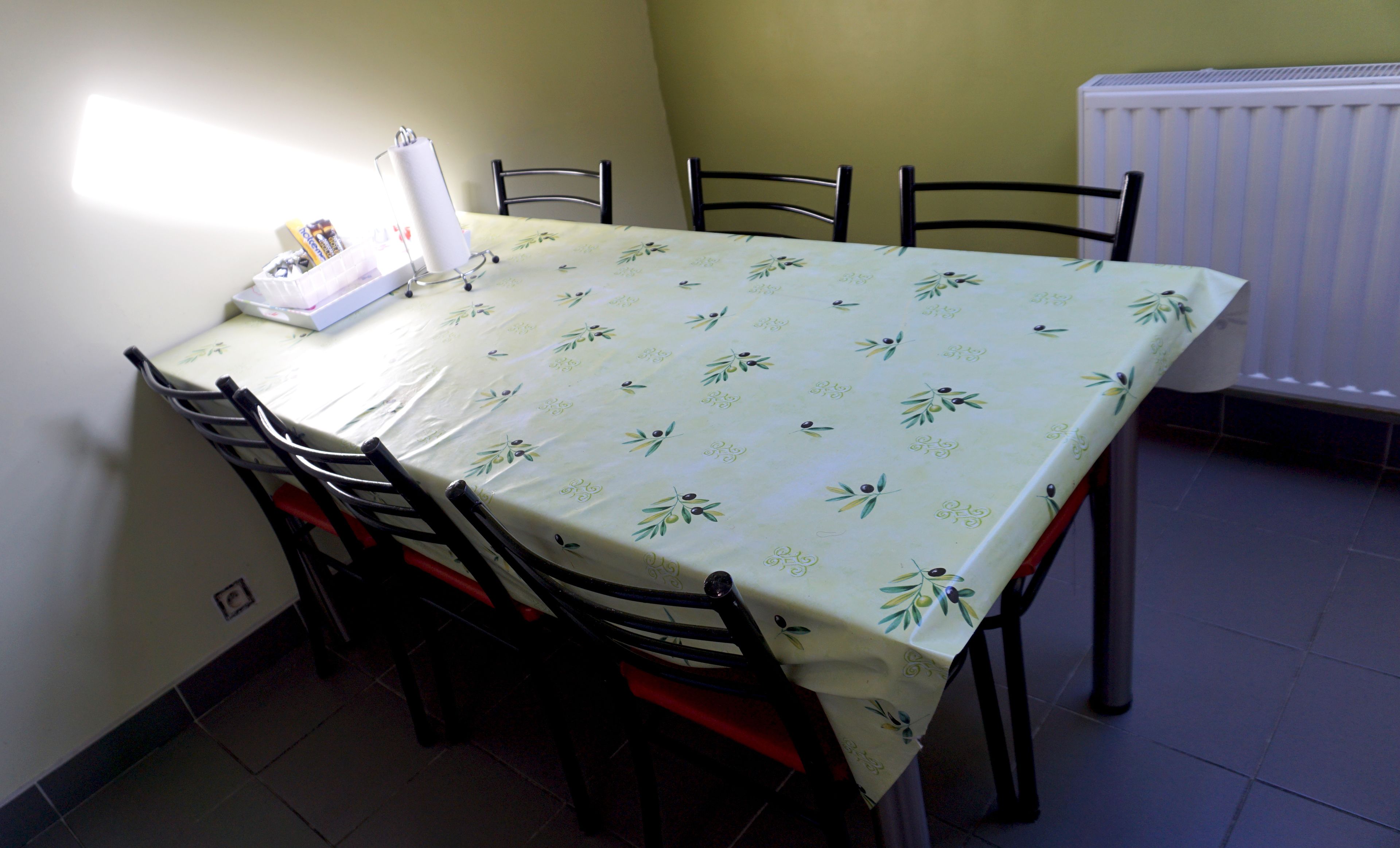 Una mesa de comedor con mantel floral, rodeada de sillas, en una habitación con radiador y una fuente de luz en la pared.