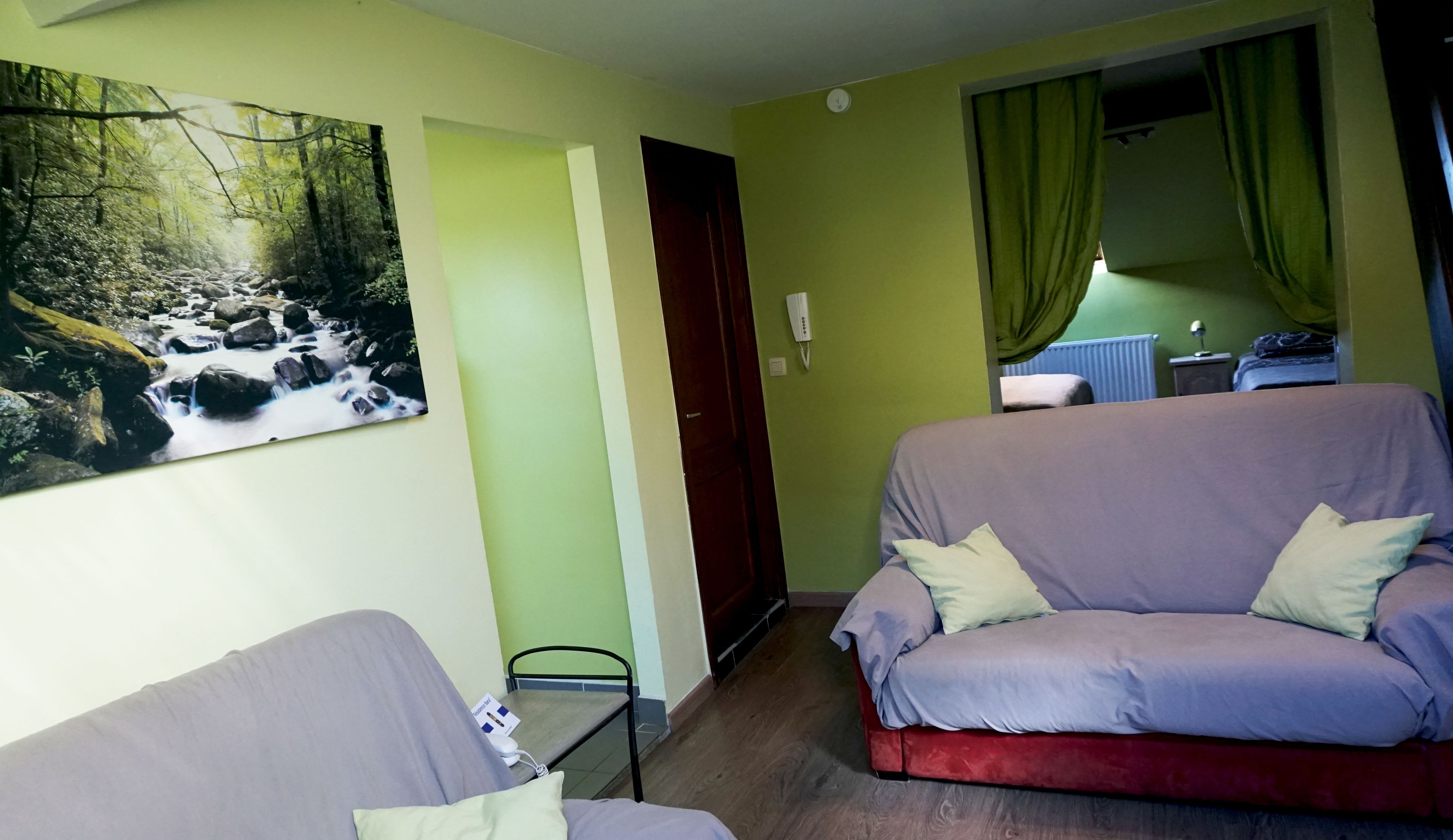 A room with a green wall, a painting of a forest stream, a red and purple sofa, a small table, and a door leading to another room.