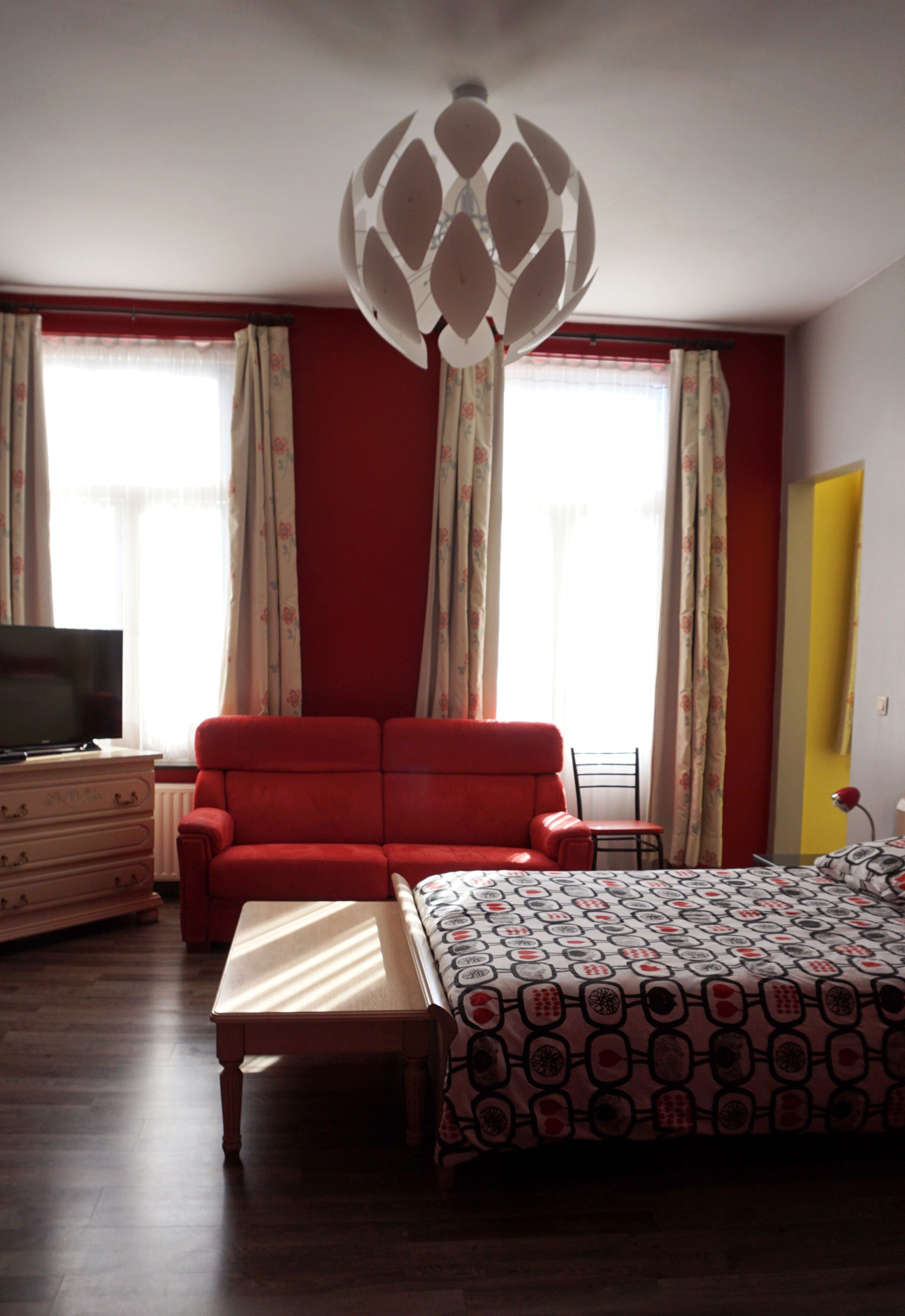 Een kamer met een rode bank, een bed met een beddeken met patroon, een houten dressoir, een televisie en een opvallende plafondlamp.