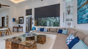 En moderne stue med et glasbord, en beige sofa med mønstrede puder og et stort vindue med udsigt til grønne omgivelser.