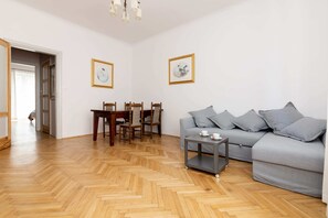 A living room with a herringbone wooden floor, a grey sofa, a small table, and a dining area with chairs and a table.