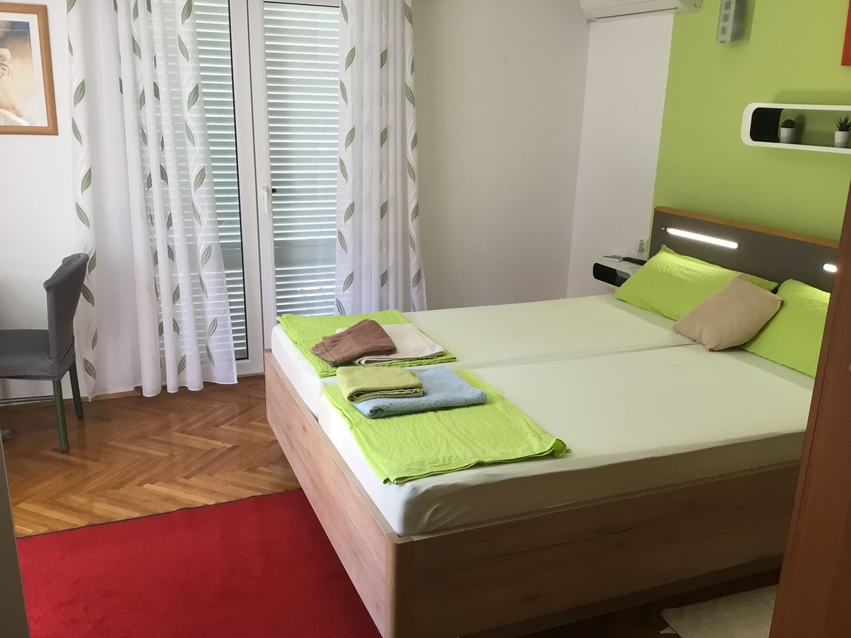 Ein ordentlich bezogenes Bett mit grünen und beigen Kissen, einem roten Teppich und einem Stuhl in einem Zimmer mit Holzboden und weißen Vorhängen mit Blattmuster.