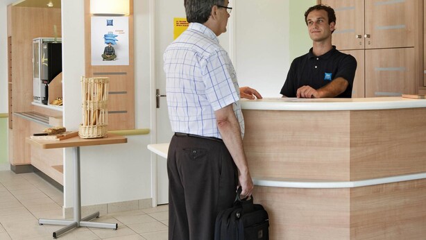 A reception area with a wooden reception desk, a counter, and a wall-mounted sign. - Ibis Budget Frankfurt Offenbach Sued (Offenbach am Main)