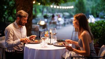Una pareja cenando en una mesa al aire libre con mantel blanco, rodeada de follaje y luces colgantes.