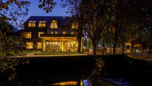 A modern two-story house with a large glass facade, a visible outdoor seating area, and a small pond in the foreground. - Sunrose 7 - Gourmet & SPA Hotel (16+) (Bohinj)