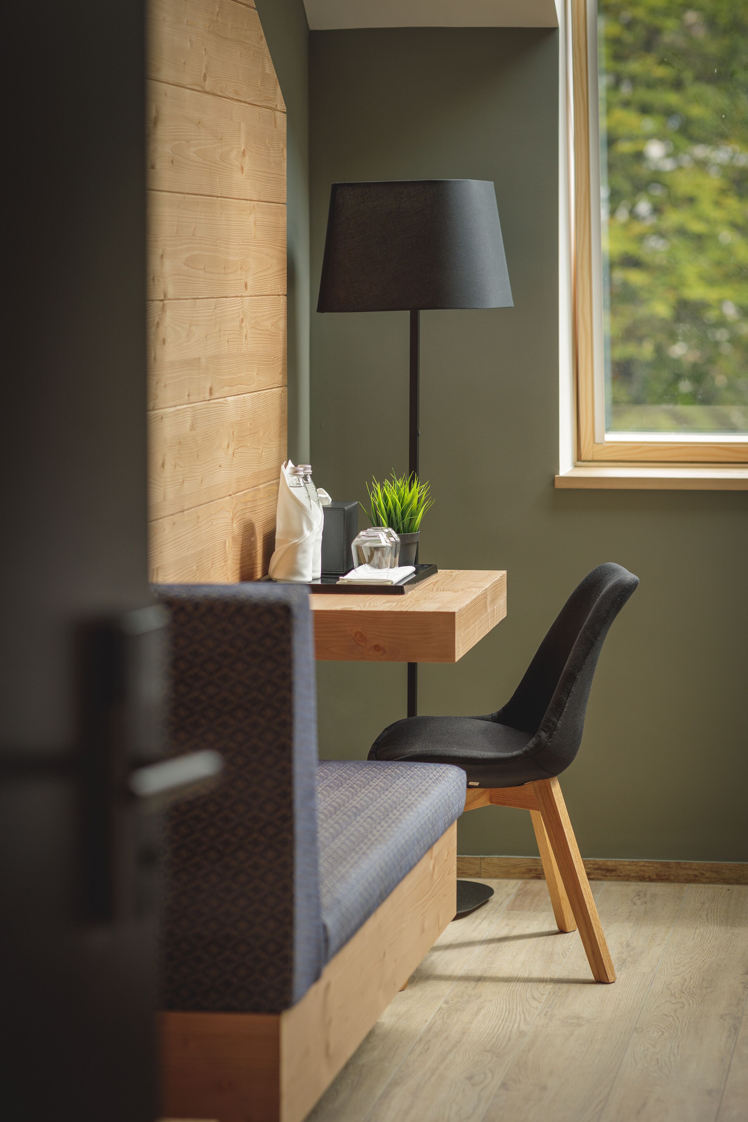 A modern room with a wooden bench, a desk with a lamp, a potted plant, and a view of greenery outside.