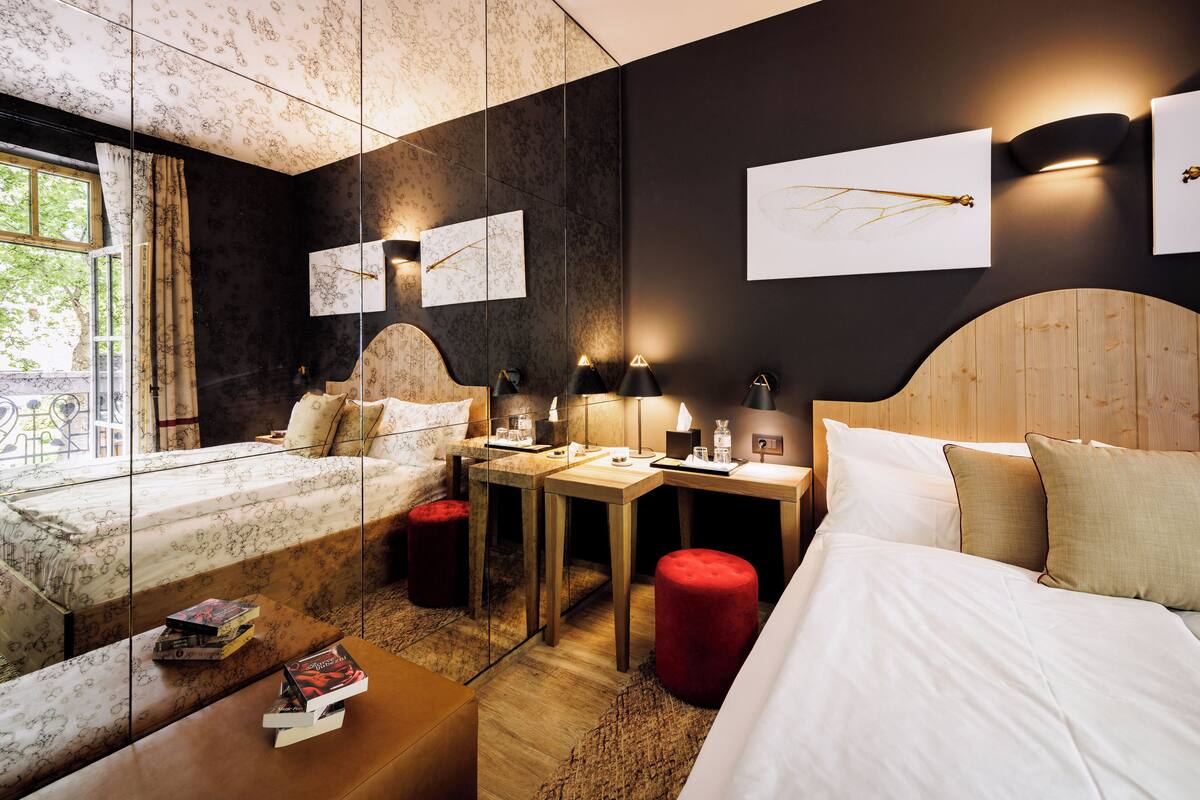 Une chambre d’hôtel comprenant un lit, un bureau, deux chaises, un pouf rouge et des œuvres d’art accrochées aux murs.