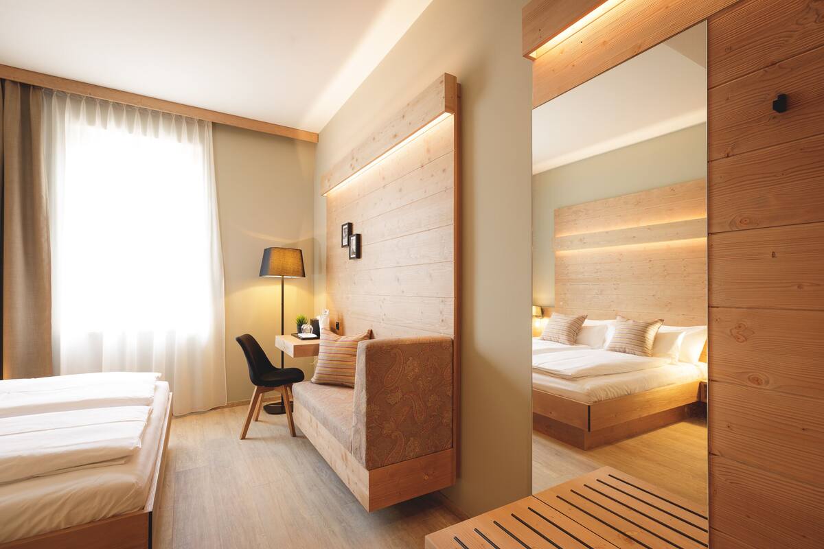 Une chambre d’hôtel moderne avec une tête de lit en bois, un canapé-lit, un bureau avec une lampe et un grand miroir.