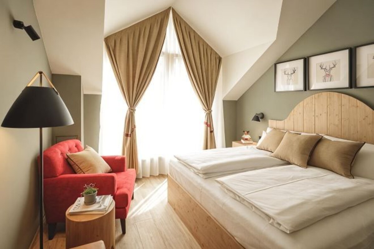Un dormitorio con una cama, una silla roja, una mesita de noche con lámpara y una ventana con cortinas.