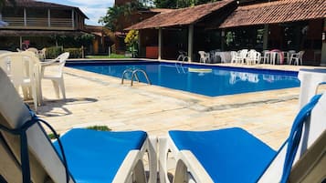 Piscina com espreguiçadeiras e mesas, cercada por edifícios e vegetação.