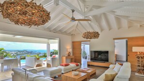 Ein großzügiges Wohnzimmer mit weißen Sofas, einem Holztisch und einem großen Fernseher. Ein Deckenventilator und ein Blick auf den Ozean durch große Fenster.