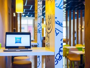 Pöydällä on kannettava tietokone, jonka näytöllä näkyy ibis budget -hotelliketjun logo. Huoneen moderni sisustus sisältää abstraktia seinätaidetta ja viihtyisiä oleskelualueita.