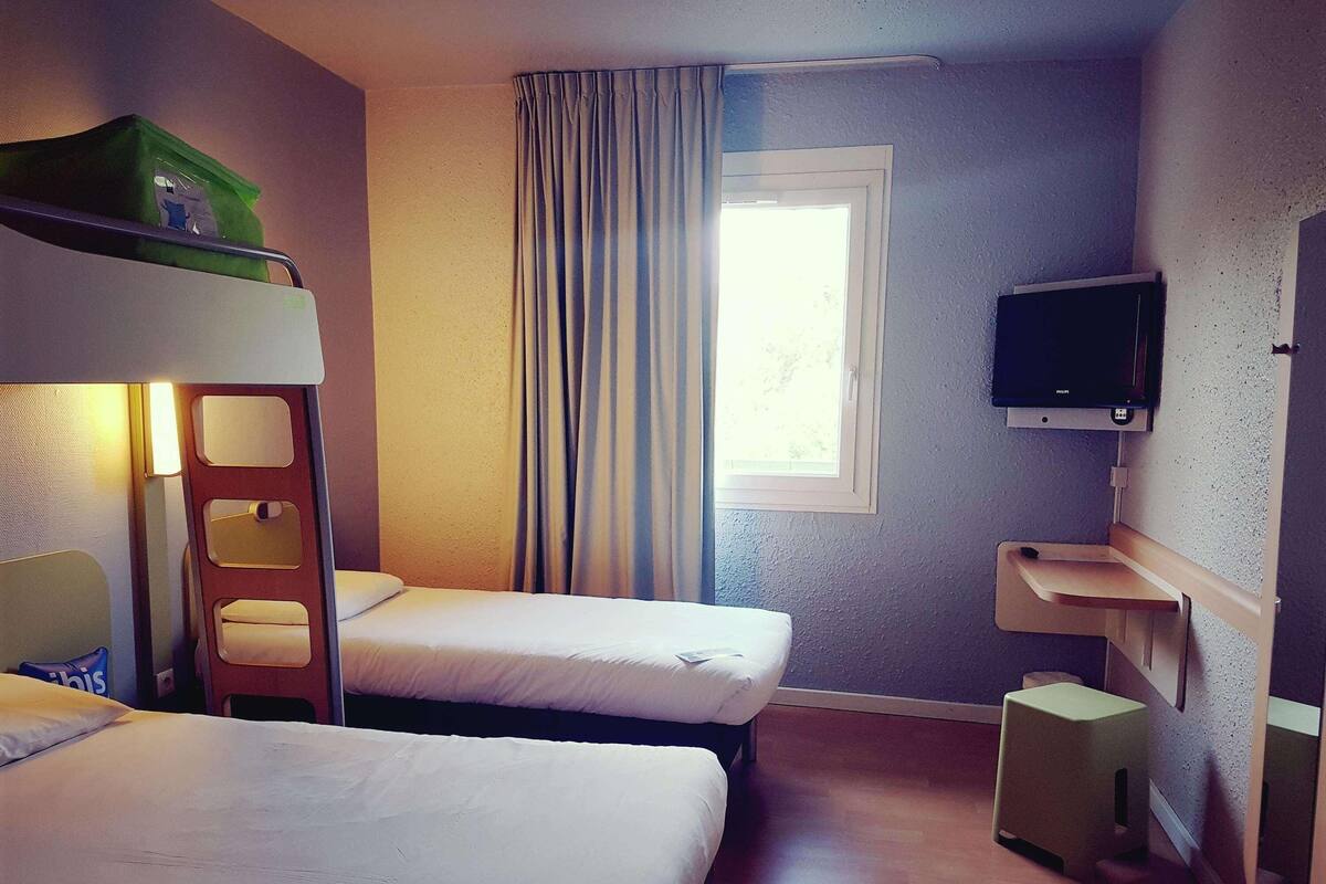Una habitación de hotel con literas, televisión, ventana con cortinas y un taburete de madera.