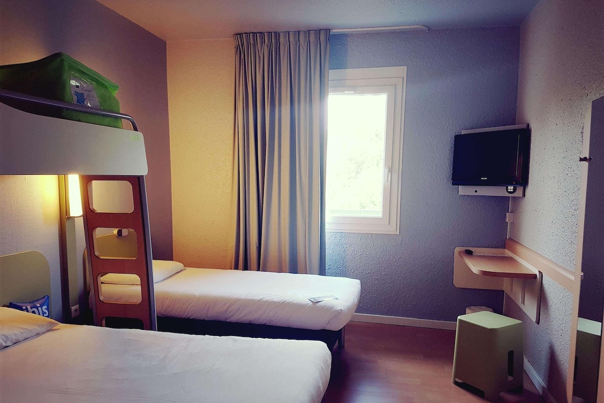 Une chambre d’hôtel avec des lits superposés, une télévision, une fenêtre avec des rideaux et un petit tabouret en bois.