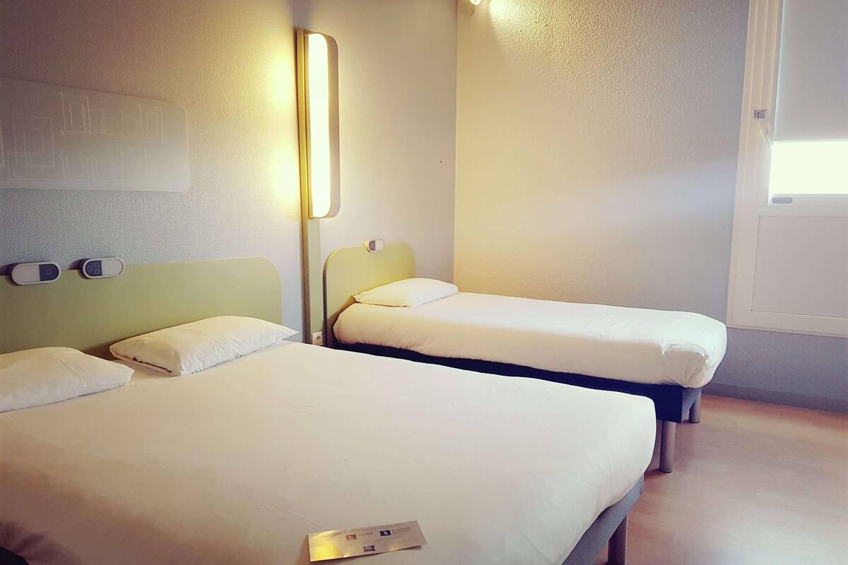 Habitación de hotel con dos camas, una mesita de noche, una lámpara de pared y un ventanal.