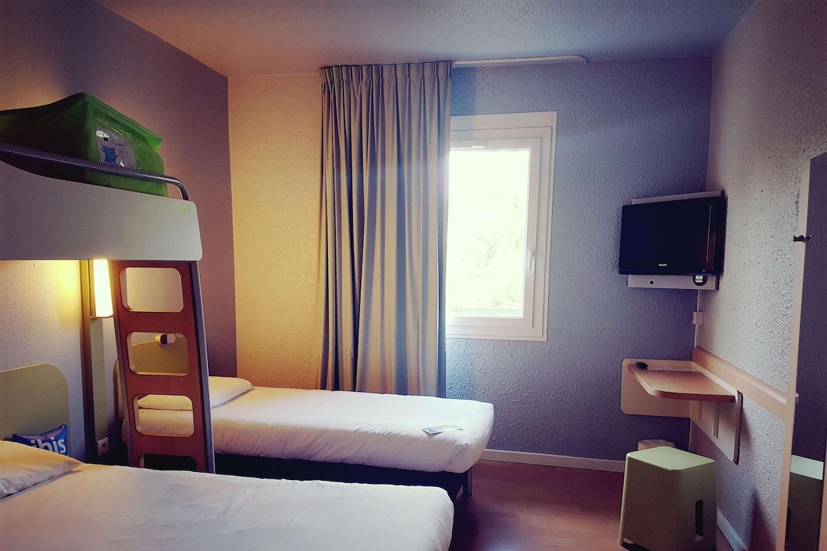Una habitación de hotel con literas, televisión, ventana con cortinas y un taburete de madera.