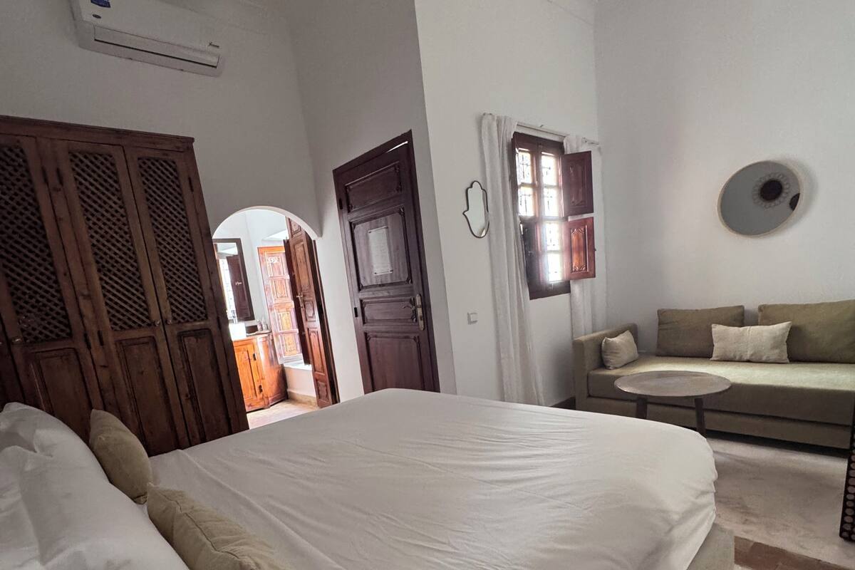 A bedroom with a large bed, a sofa, and a small table. There is a door leading to another room and a window with curtains.