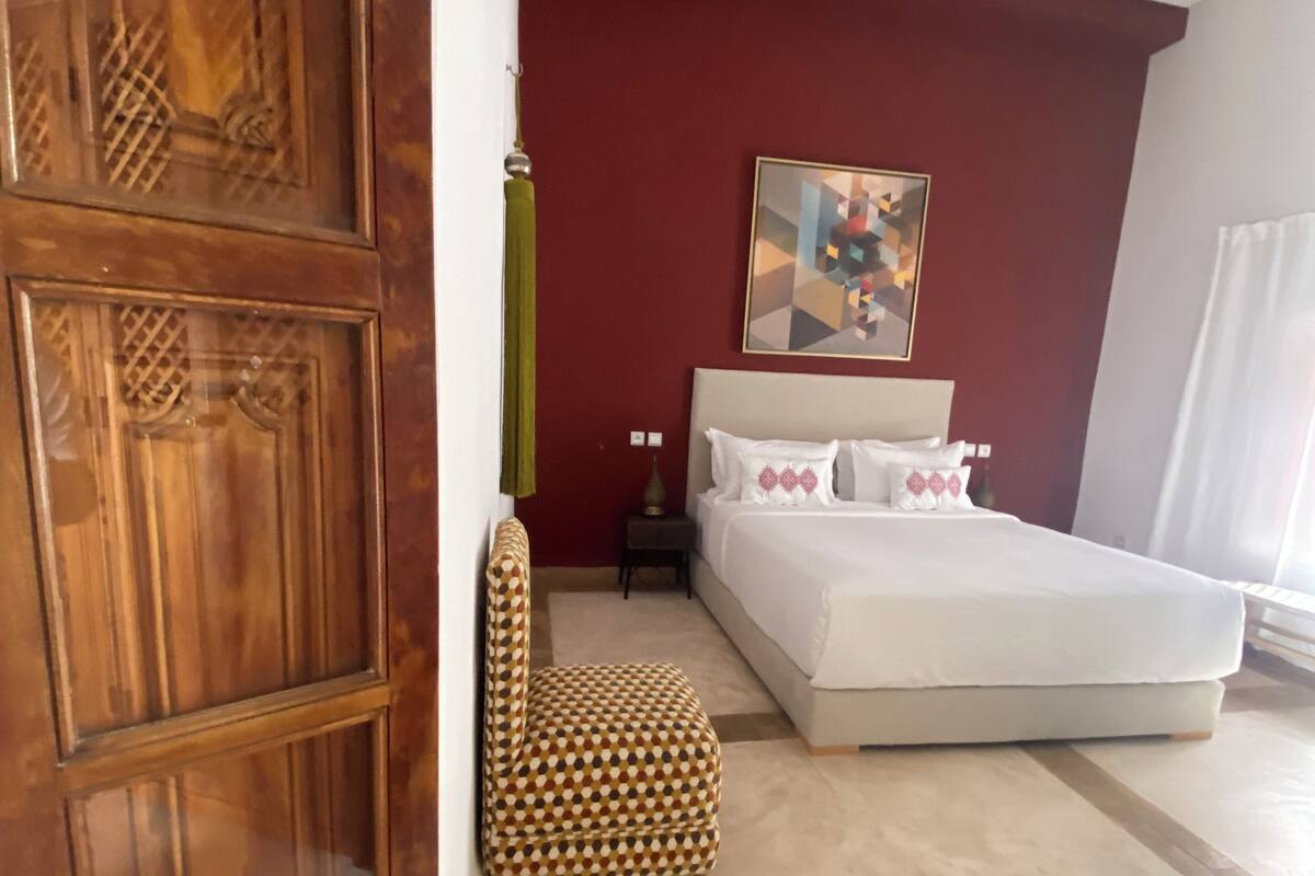 A bedroom with a wooden door, a bed with white bedding, a patterned armchair, and a painting on the wall.