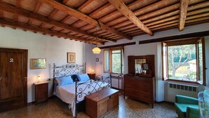 Un dormitorio con techo de madera, cama con estructura metálica, cómoda, espejo y ventana con vista a árboles.