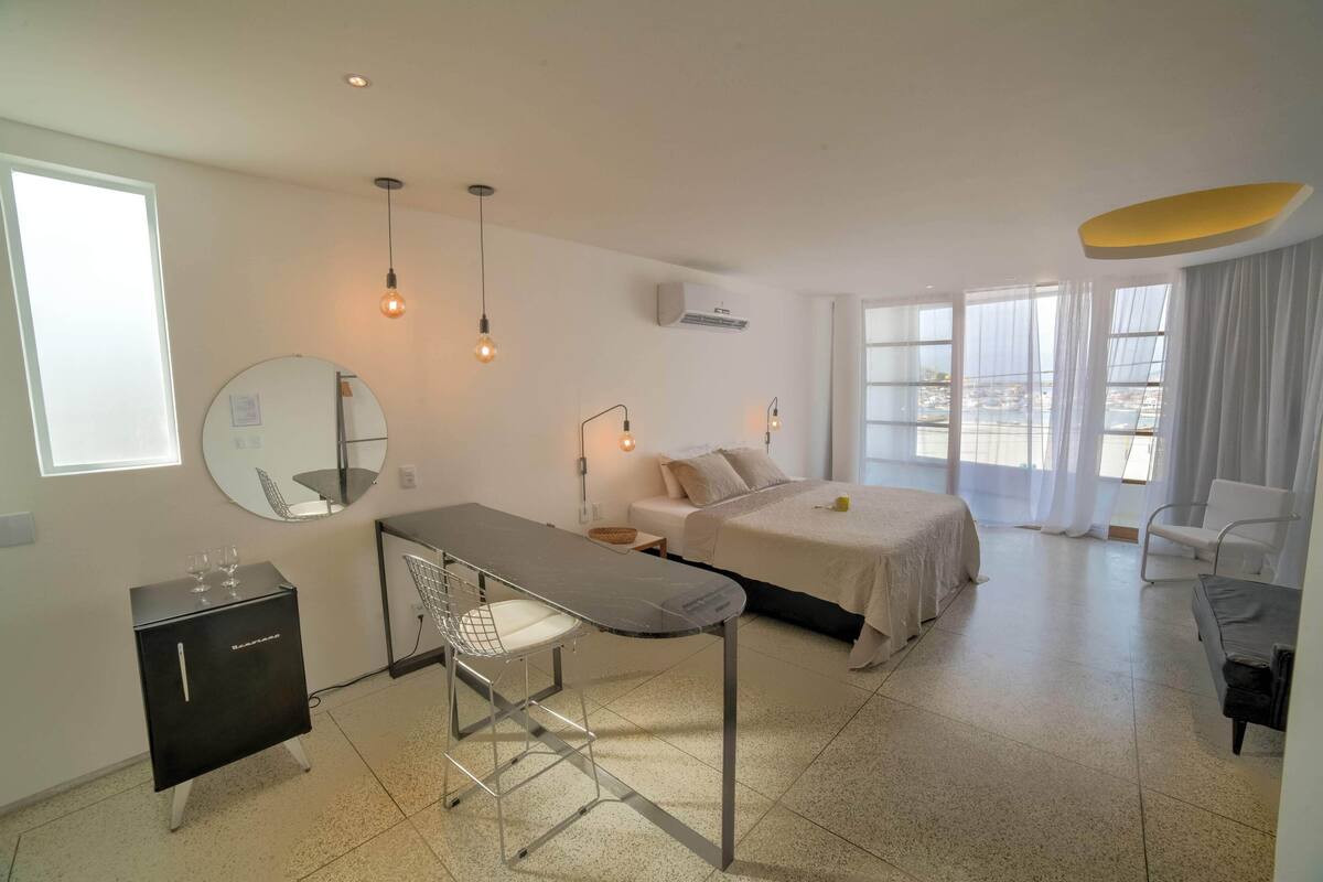 A modern bedroom with a bed, bedside table, and a desk with a chair. There is a window with curtains, a mirror, and a hanging light fixture.