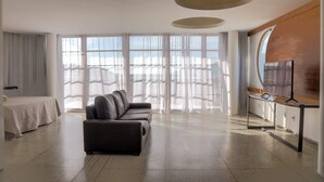 Una habitación de hotel moderna con una cama grande, un sofá, un televisor y ventanas amplias con cortinas translúcidas.