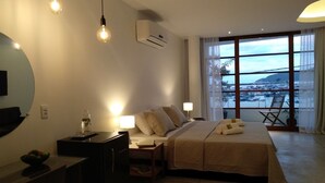 Habitación de hotel con cama, mesitas de noche, escritorio, silla, televisor y un amplio ventanal con vistas al mar.