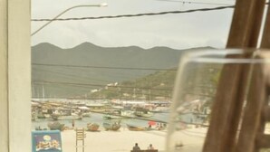 Vista desde una ventana a un puerto deportivo con barcos, cielo despejado y una copa de vino blanco sobre una mesa.