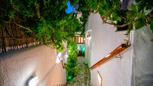 A narrow stone pathway flanked by white walls and lush greenery, leading to a building entrance. - Family Room (4-5 Adults) - Semi Basement, near Center, Parikia (Paros Greece)