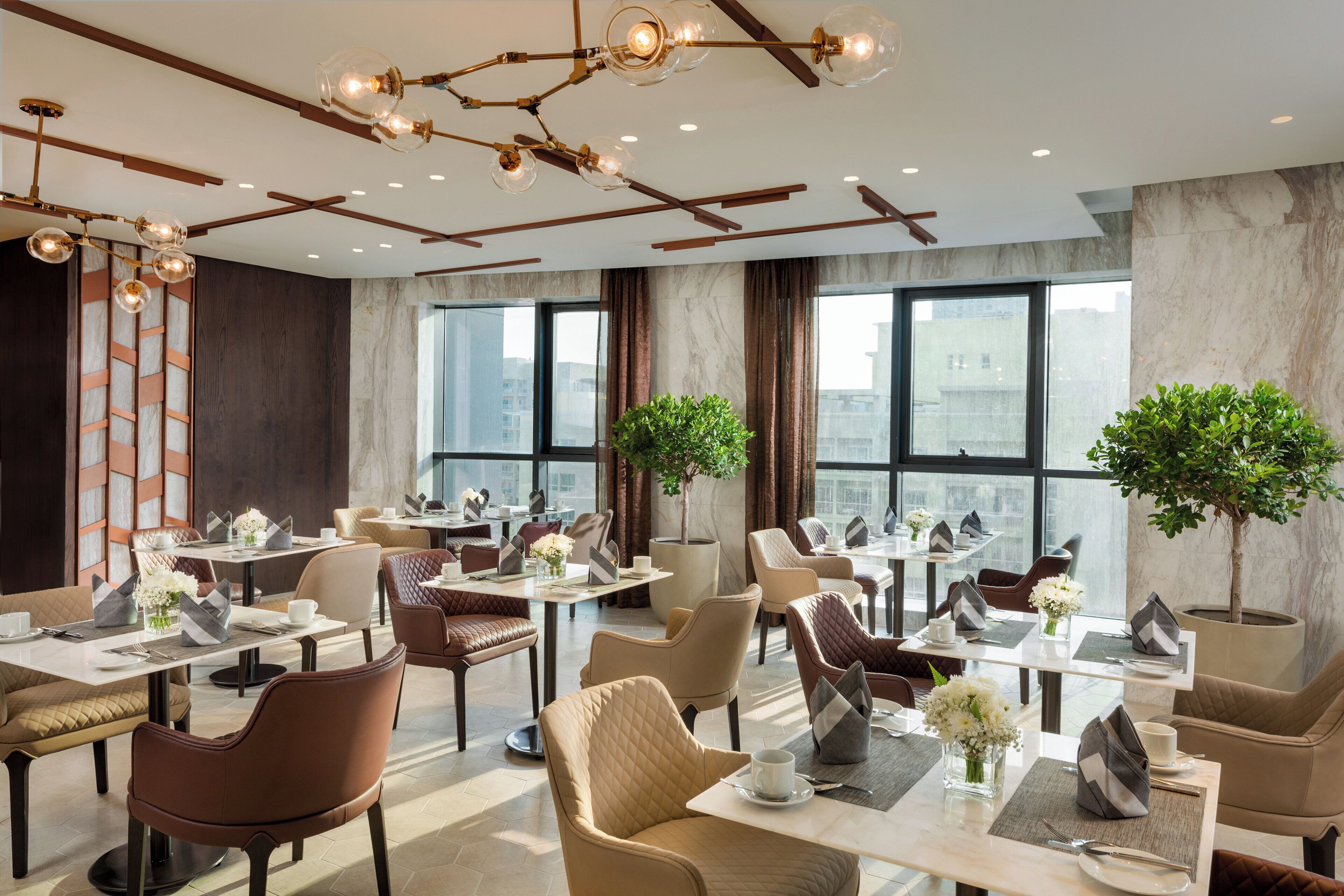 A modern hotel dining area with multiple tables and chairs, large windows, and decorative lighting.