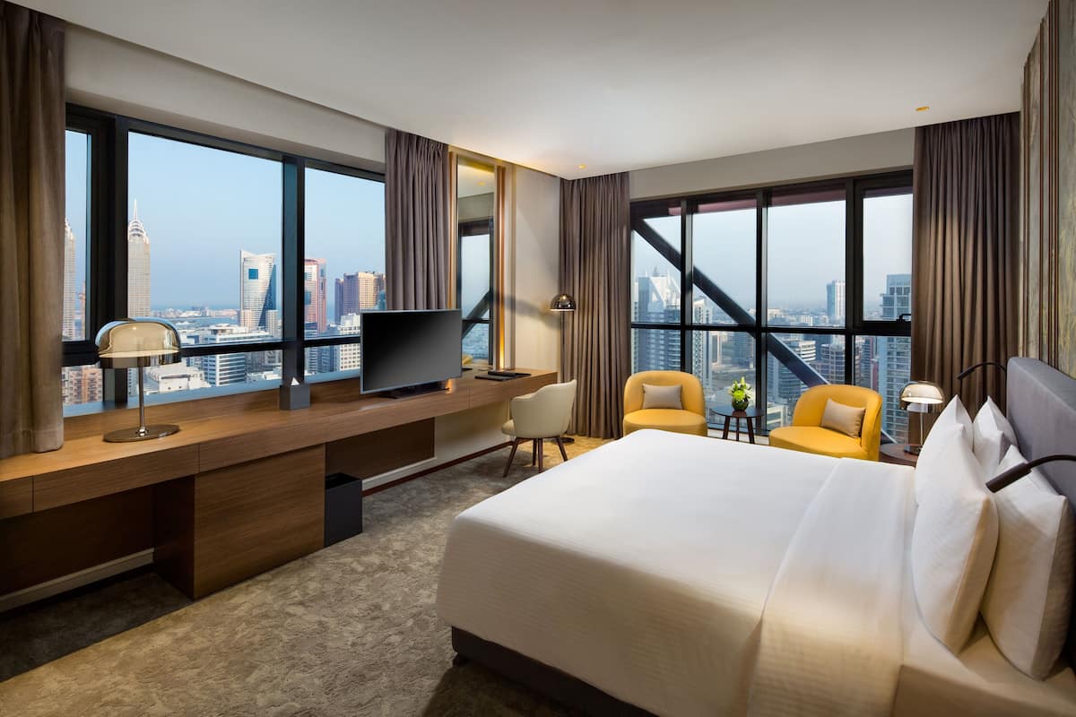 Habitación de hotel con una cama grande, un escritorio con televisor, dos sillones y vistas a la silueta urbana a través de amplias ventanas.