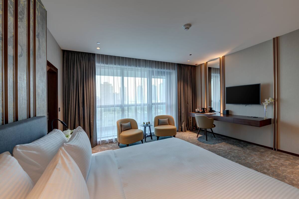 Habitación de hotel con una cama grande, dos sillones, un escritorio, un televisor y un ventanal con cortinas.