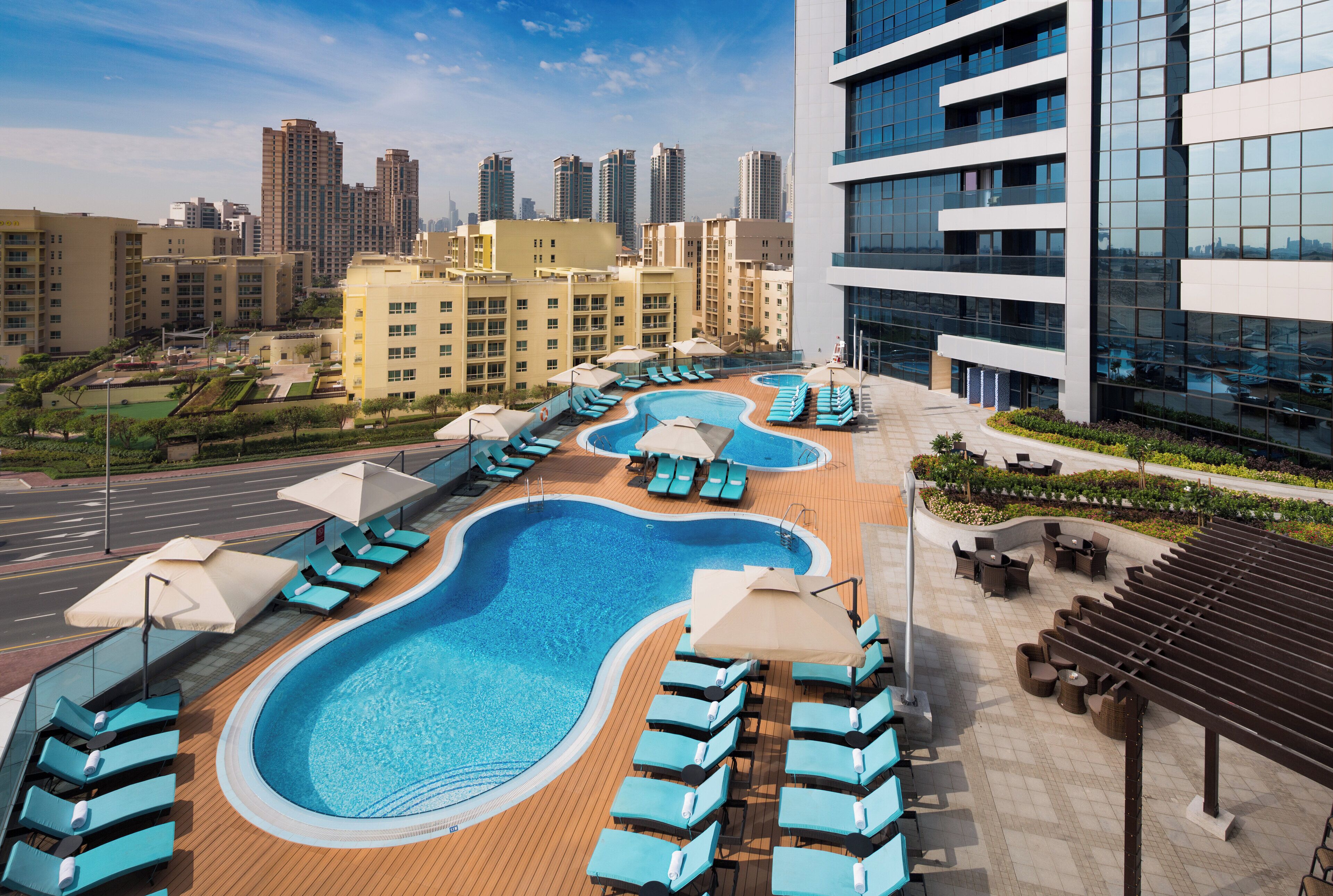 A rooftop swimming pool with lounge chairs and umbrellas, surrounded by modern buildings and a cityscape.