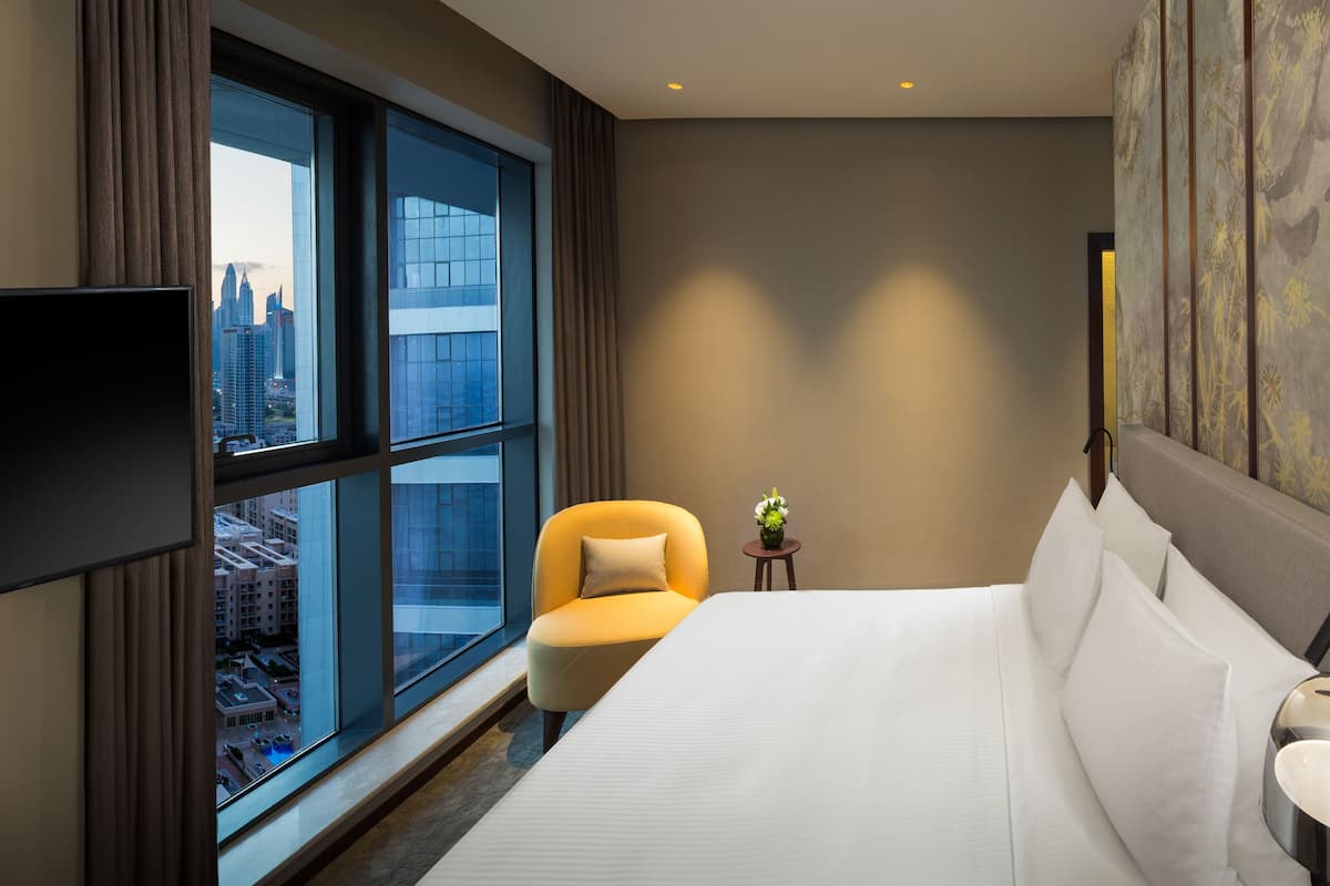 Habitación de hotel con una cama grande, un televisor montado en la pared, una ventana con vistas a la ciudad y una pequeña mesa con un jarrón de flores.