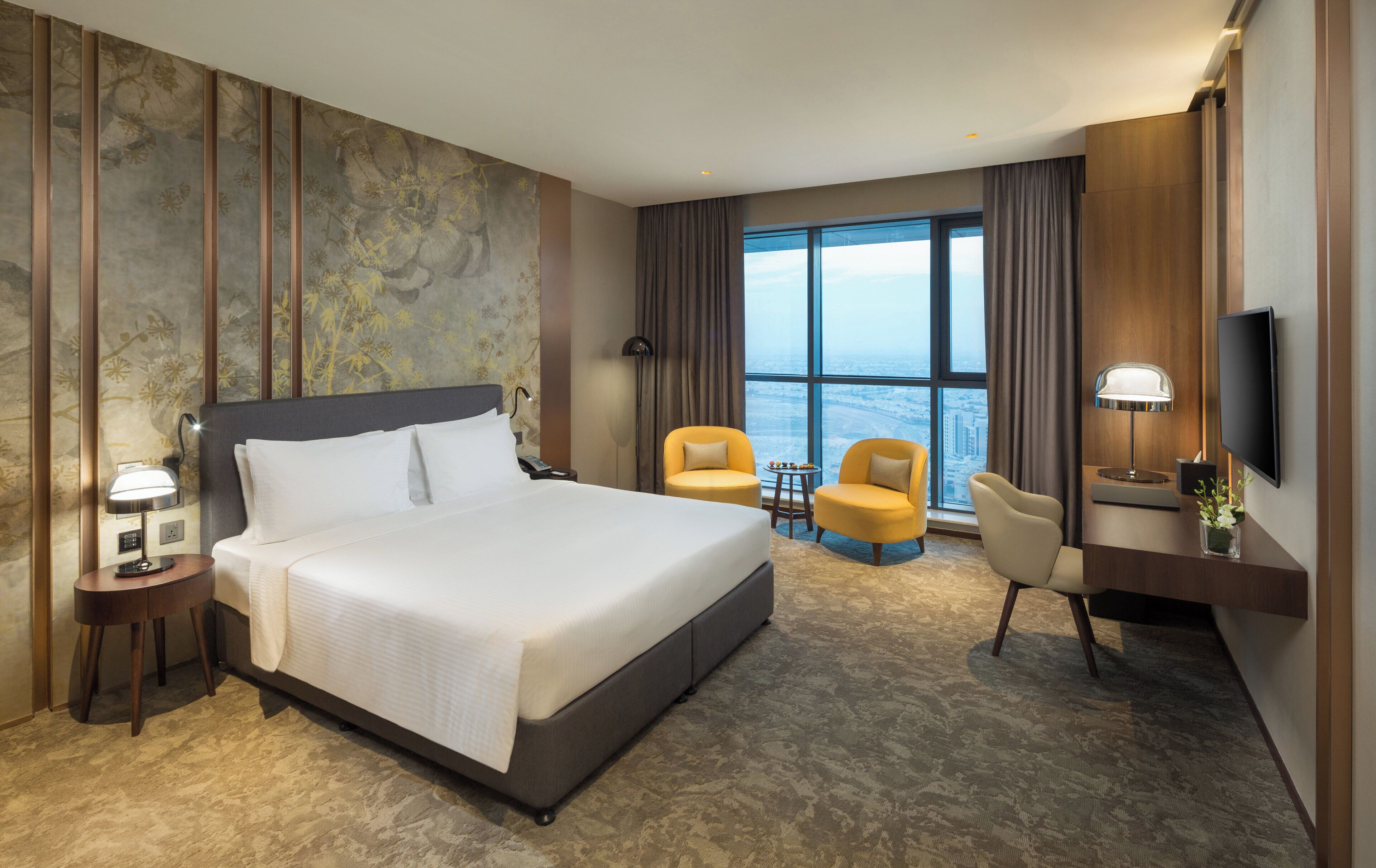 Une chambre d’hôtel avec un grand lit, deux fauteuils, un bureau avec une télévision et une vue sur la ville grâce à de grandes fenêtres.