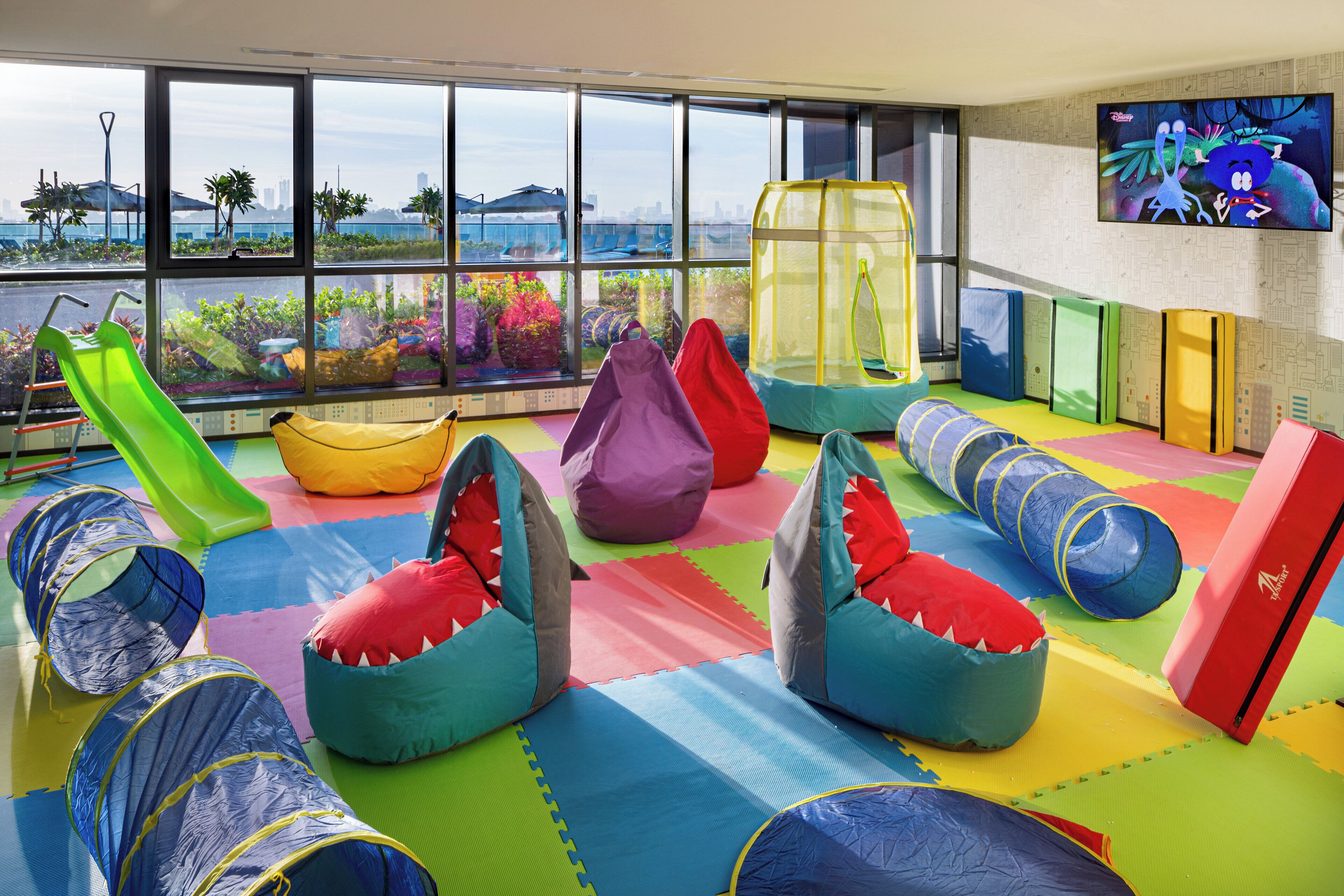 A children's play area with various play structures, including slides, tunnels, and bean bags, set on colorful mats with a large window offering a view of the city.