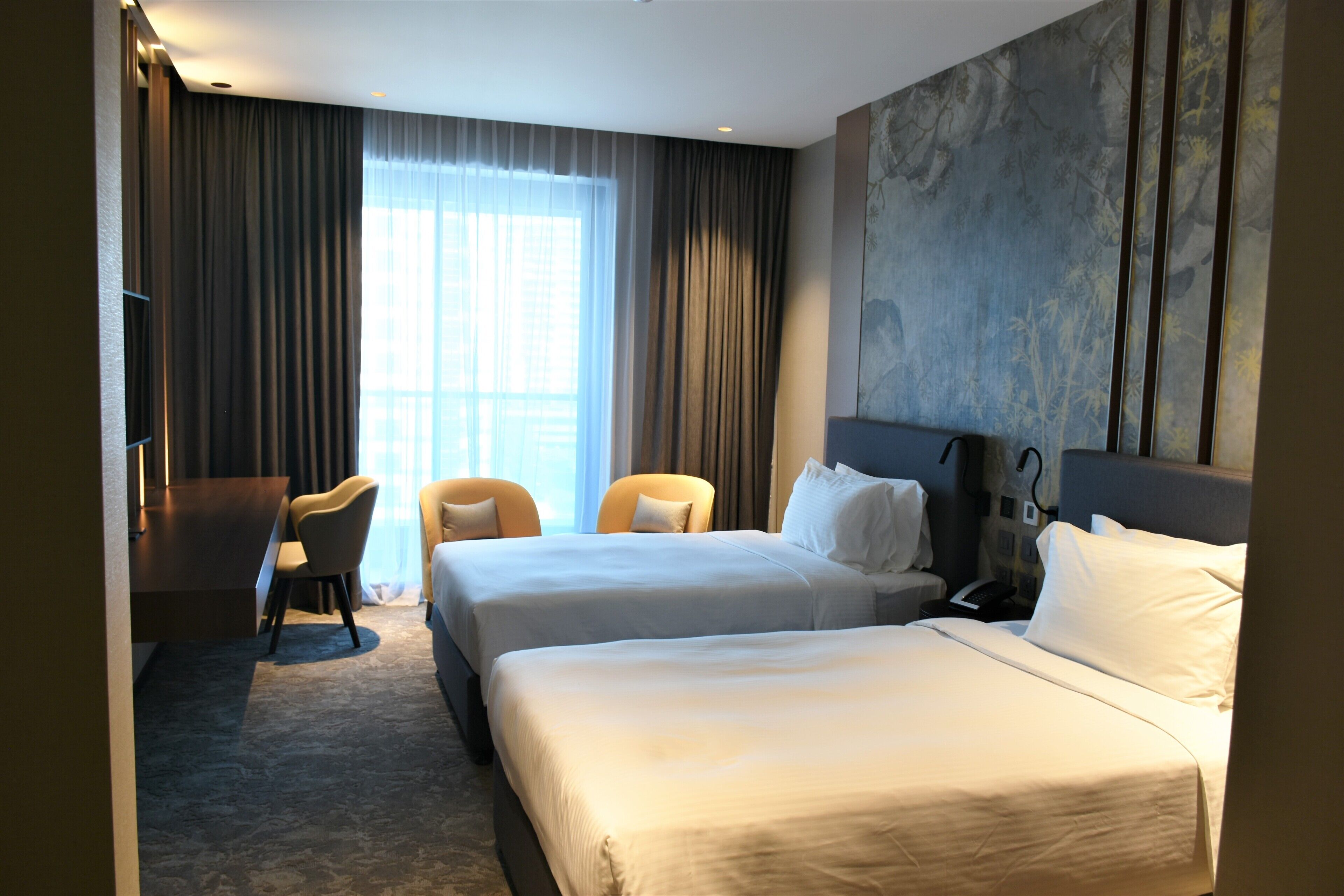 A hotel room with two beds, a desk, a chair, and a large window with curtains.