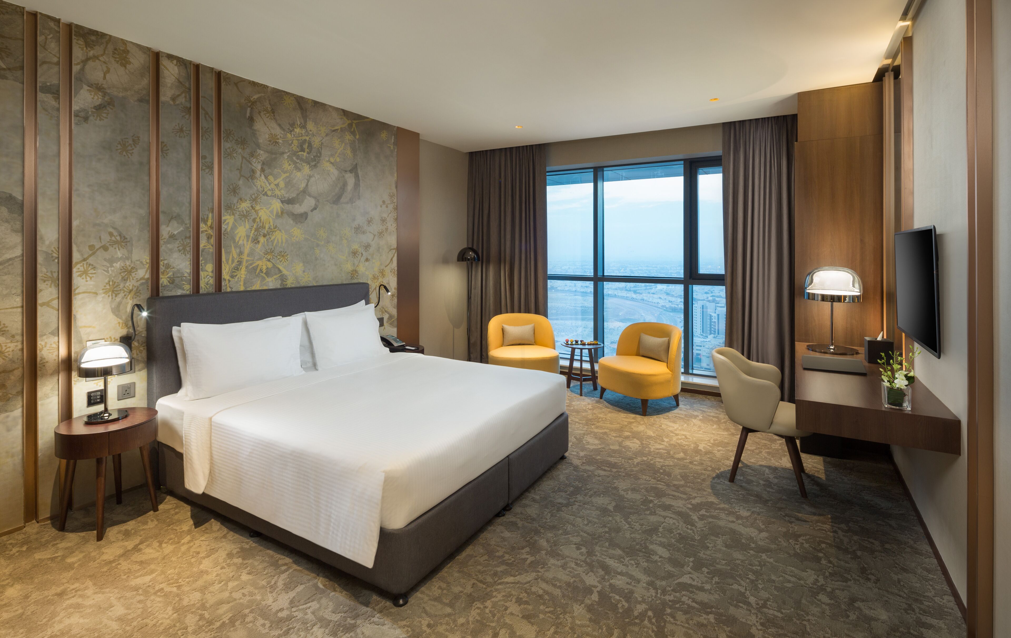 A hotel room with a large bed, two chairs, a desk with a TV, and a view of the city through large windows.