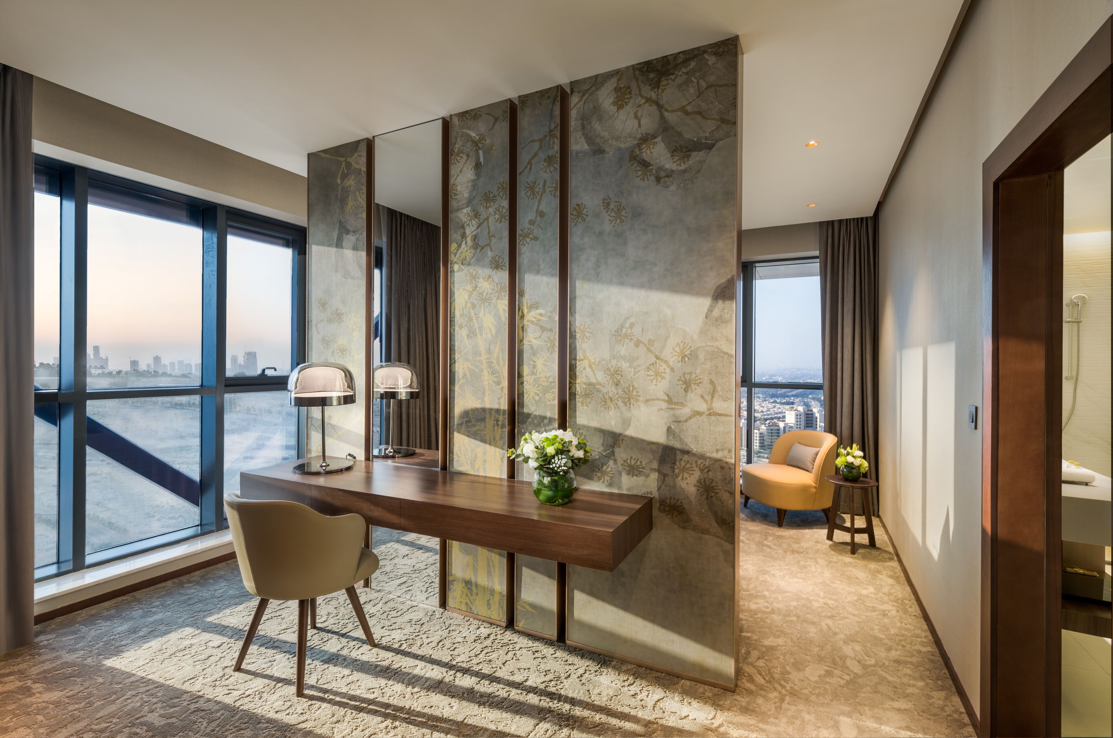 A modern hotel room with a large window, a wooden desk, a chair, and a decorative vase.