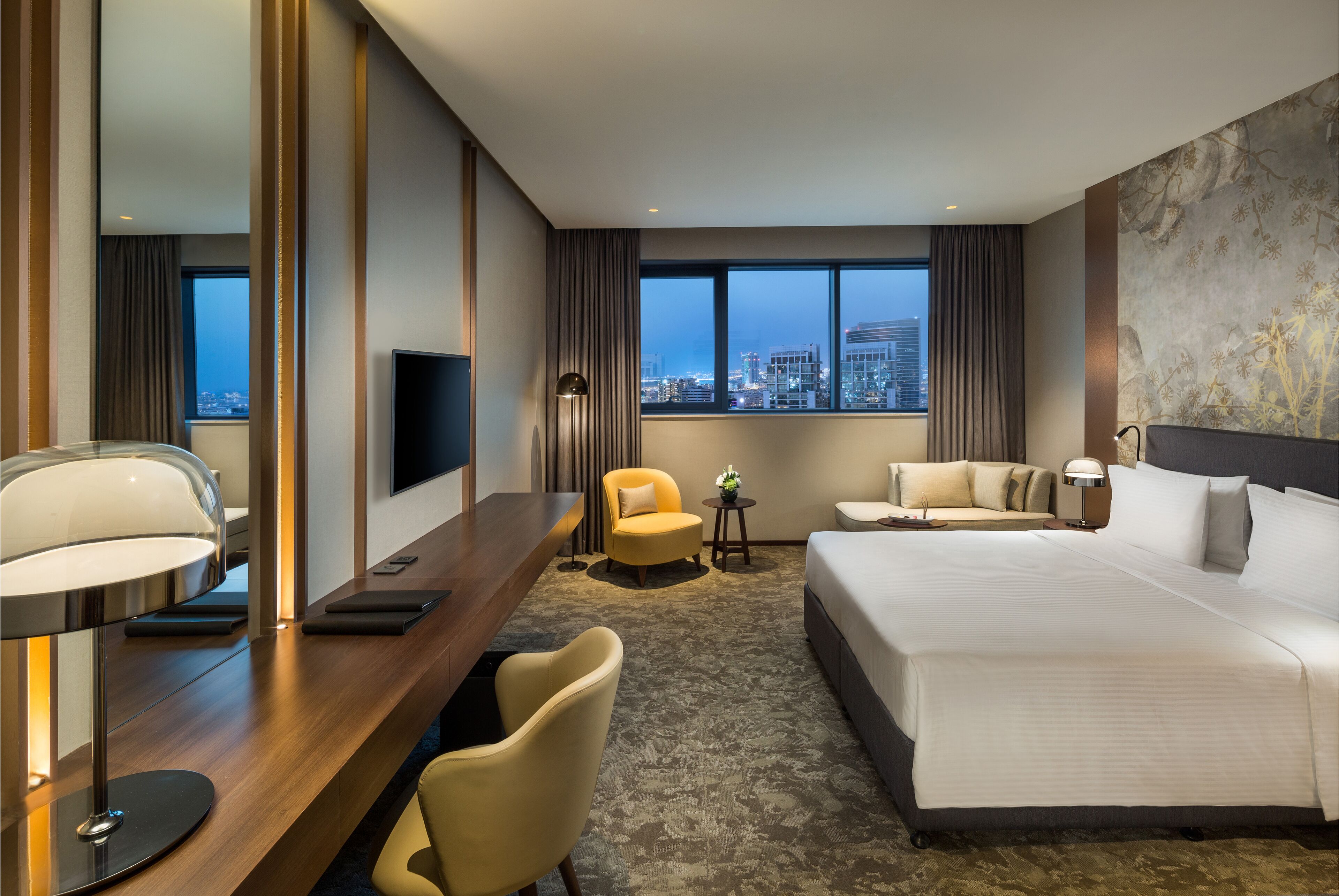 Ein modernes Hotelzimmer mit einem großen Bett, einem Schreibtisch mit Lampe, einem Sessel, einer Couch, einem Fernseher und Blick auf die Stadtlandschaft.
