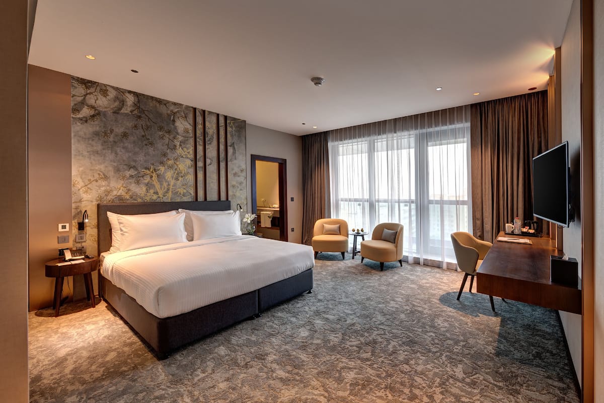 Una habitación de hotel moderna con una cama grande, dos sillones, un escritorio con televisor y un amplio ventanal con cortinas.
