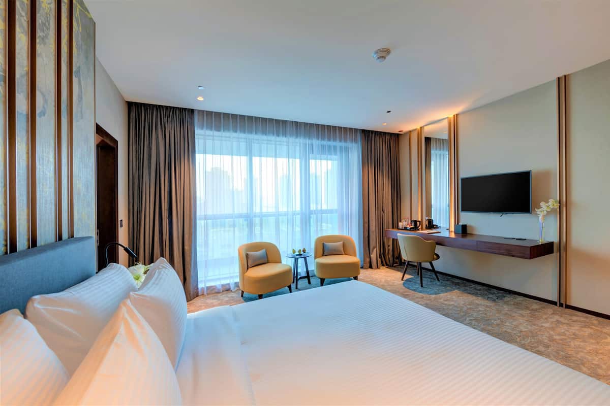 Una habitación de hotel con una cama grande, dos sillones, un escritorio, un televisor de pantalla plana y un ventanal con cortinas.