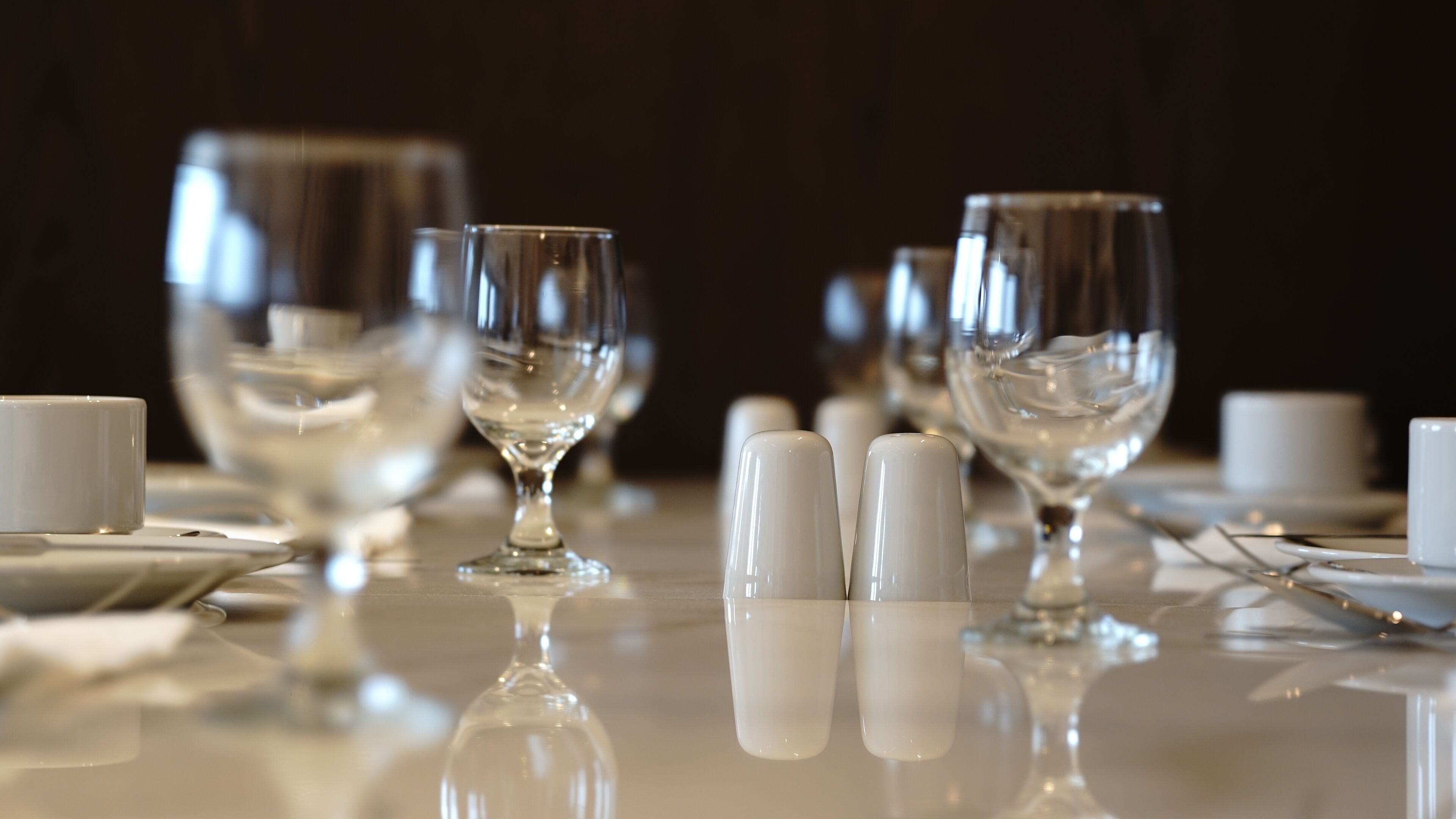 A table set with clear glassware and white cups, reflecting on a glossy surface.