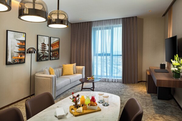 Habitación de hotel con una mesa de comedor dispuesta para el desayuno, un sofá, un televisor y obras de arte en las paredes.