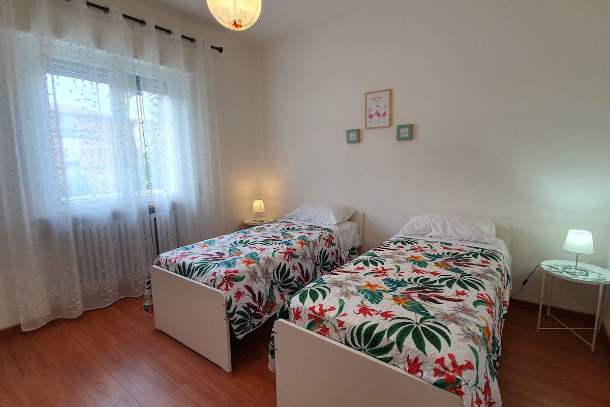 Ein Schlafzimmer mit zwei Einzelbetten, einem Fenster mit leichtem Vorhang, einem kleinen Nachttisch mit Lampe und einem an der Wand hängenden Bild.