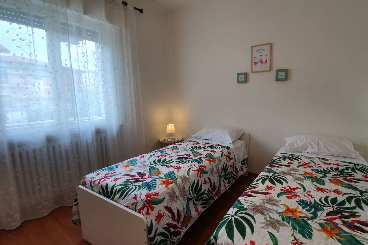 Ein Zimmer mit zwei Einzelbetten, jeweils mit einer blumigen Tagesdecke, einem Nachttisch mit Lampe und einem Fenster mit durchscheinenden Vorhängen.