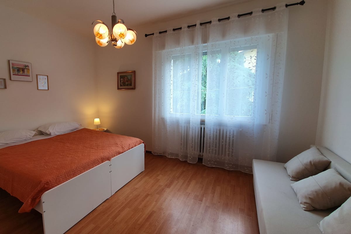 Ein Schlafzimmer mit Bett, Nachttisch mit Lampe, Fenster mit leichtem Vorhang und Holzboden.