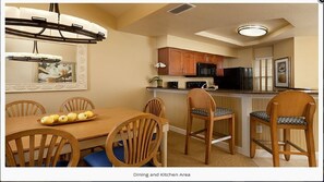 Un comedor y cocina con sillas de madera, una mesa de madera con frutero, una lámpara colgante, armarios de madera, un refrigerador negro y un microondas.